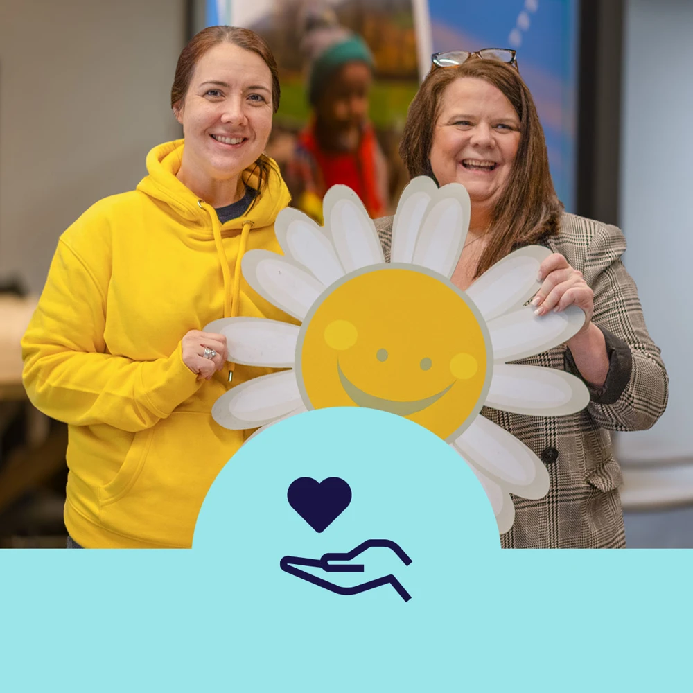 Two people posing for a photo, holding a sunflower prop.  A logo is displayed below with an icon of an outstretched hand, with a floating heart.
