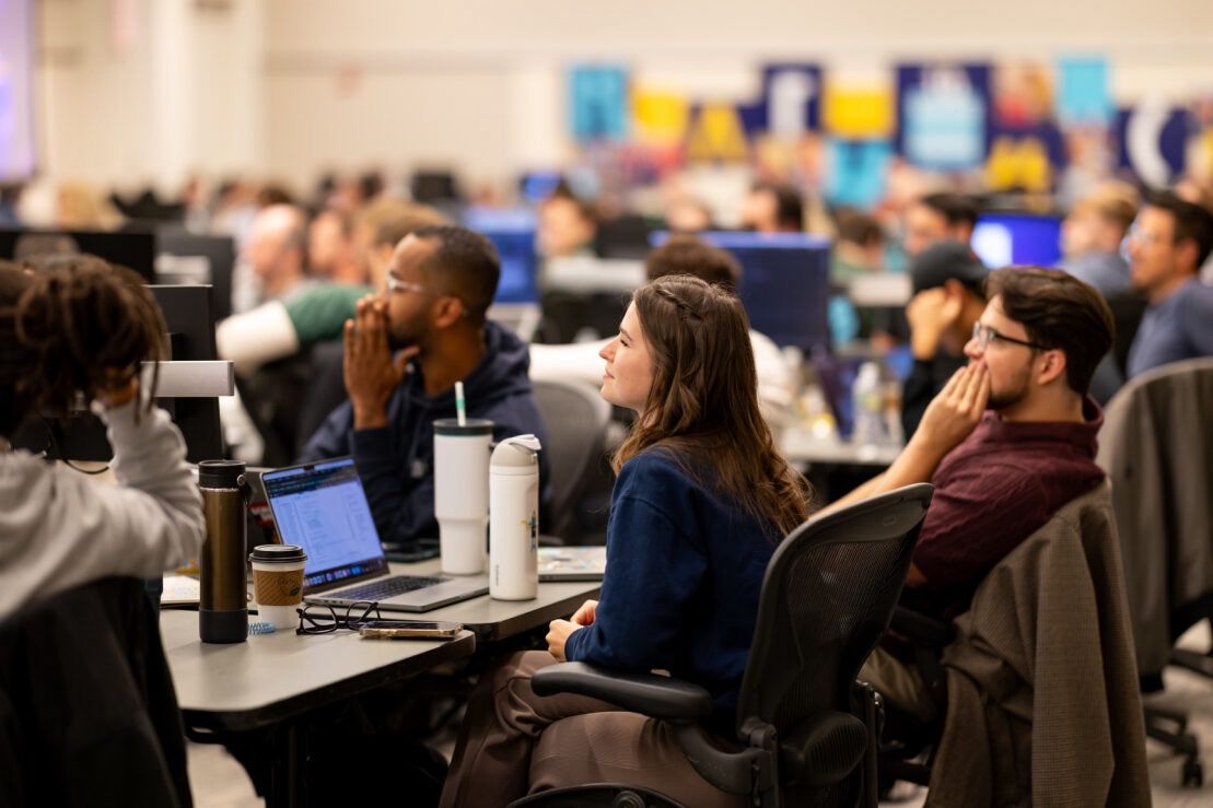 A group of people seated at desks in a conference room, attentively listening or watching something out of view. The background has blurred signage with colorful elements. Laptops and beverages are on the desks.