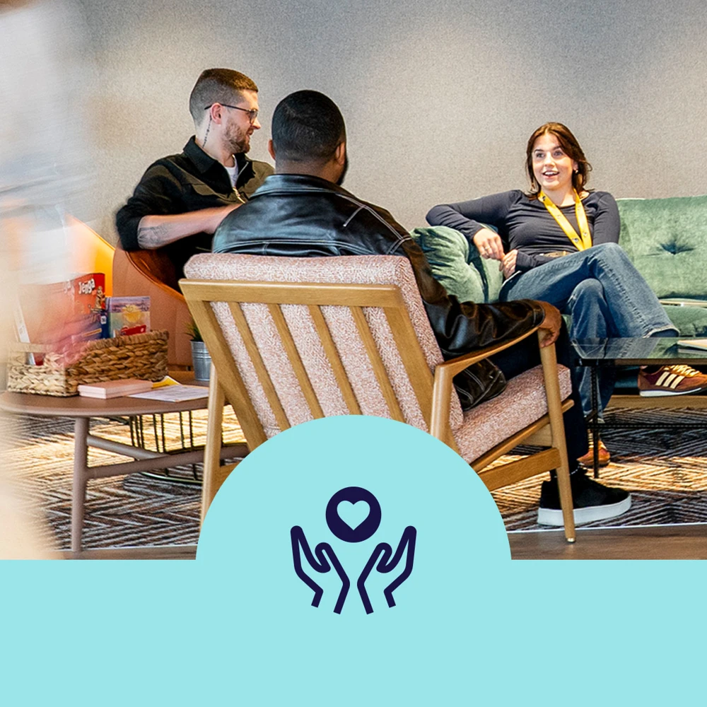 A group of people seated in a casual lounge area, featuring a wooden table with various items. The background includes a green sofa and soft lighting. A logo with two hands holding a heart is displayed at the bottom center.