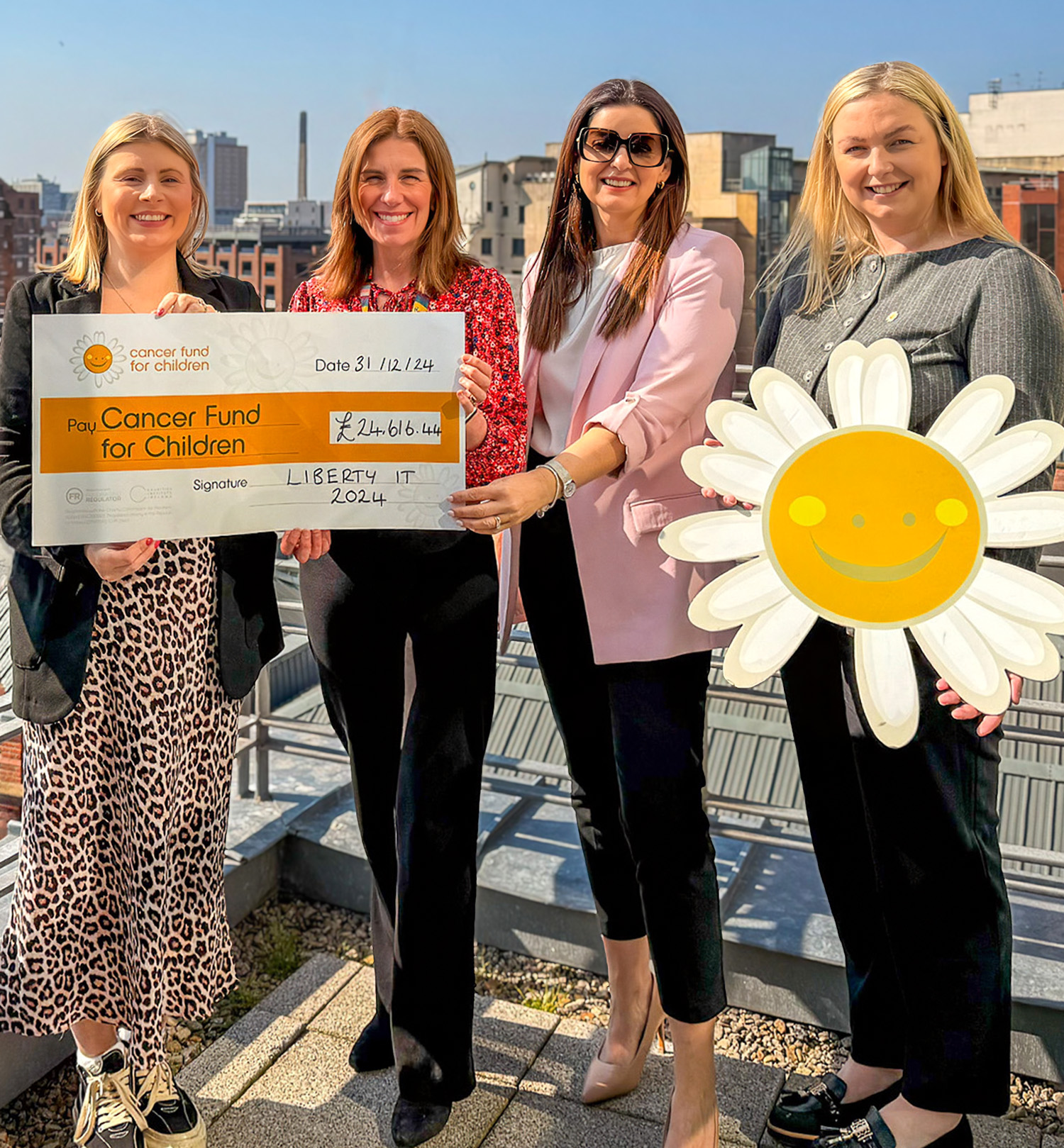 Four individuals standing on a rooftop terrace, holding a large check for Cancer Fund for Children and a cheerful sunflower sign. The donation amount is £24,616.00, with "Liberty IT" mentioned. The cityscape is visible in the background.