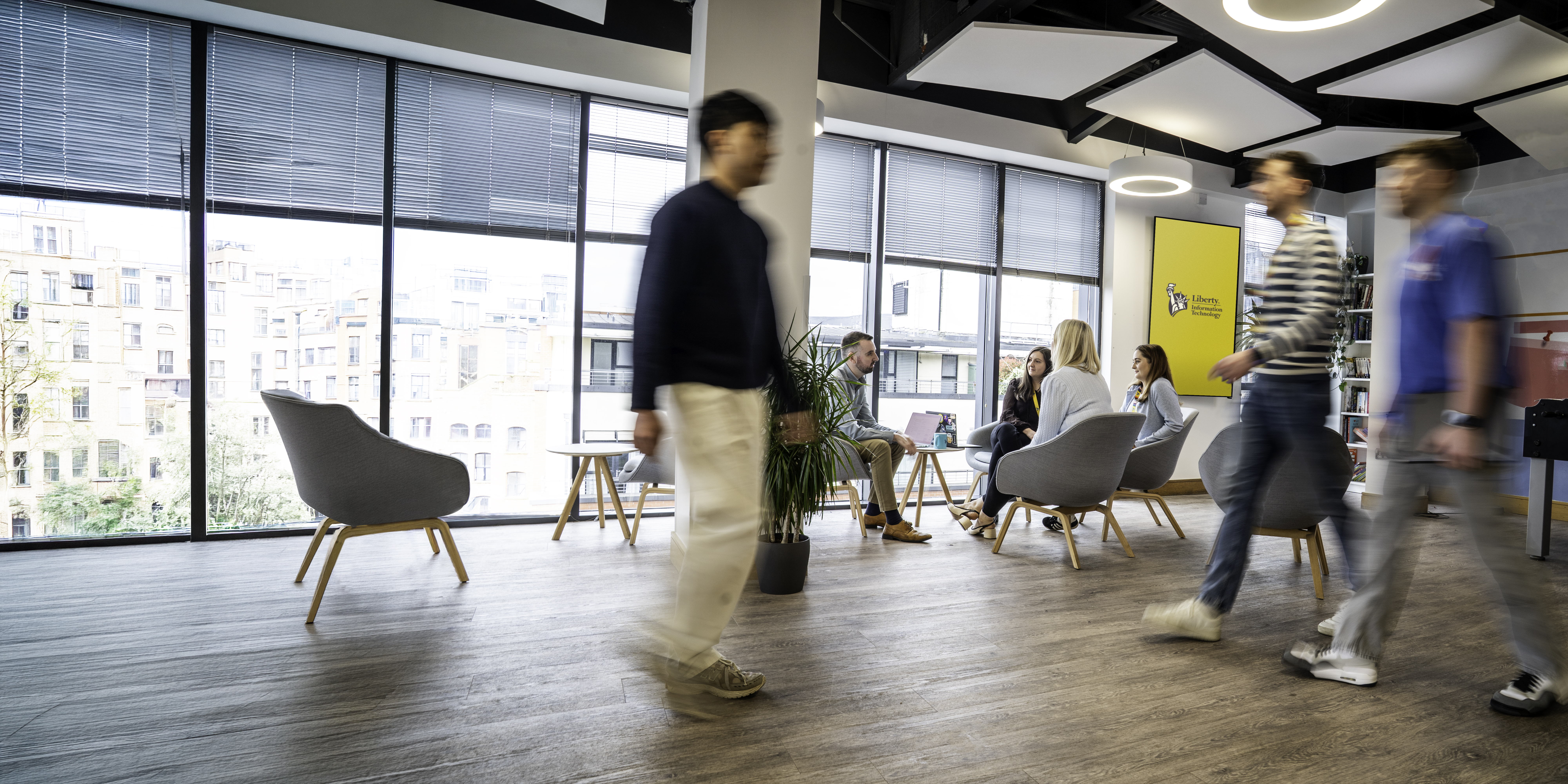 Employees walking through a bright Liberty IT office space while colleagues meet in a seating area.