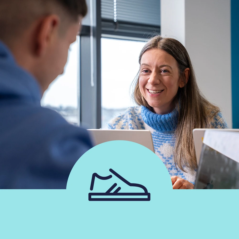 Two people are sitting at a table with laptops open in an office setting. One person is smiling at the other. There's a graphic of a shoe on a blue bar in the lower part of the image.