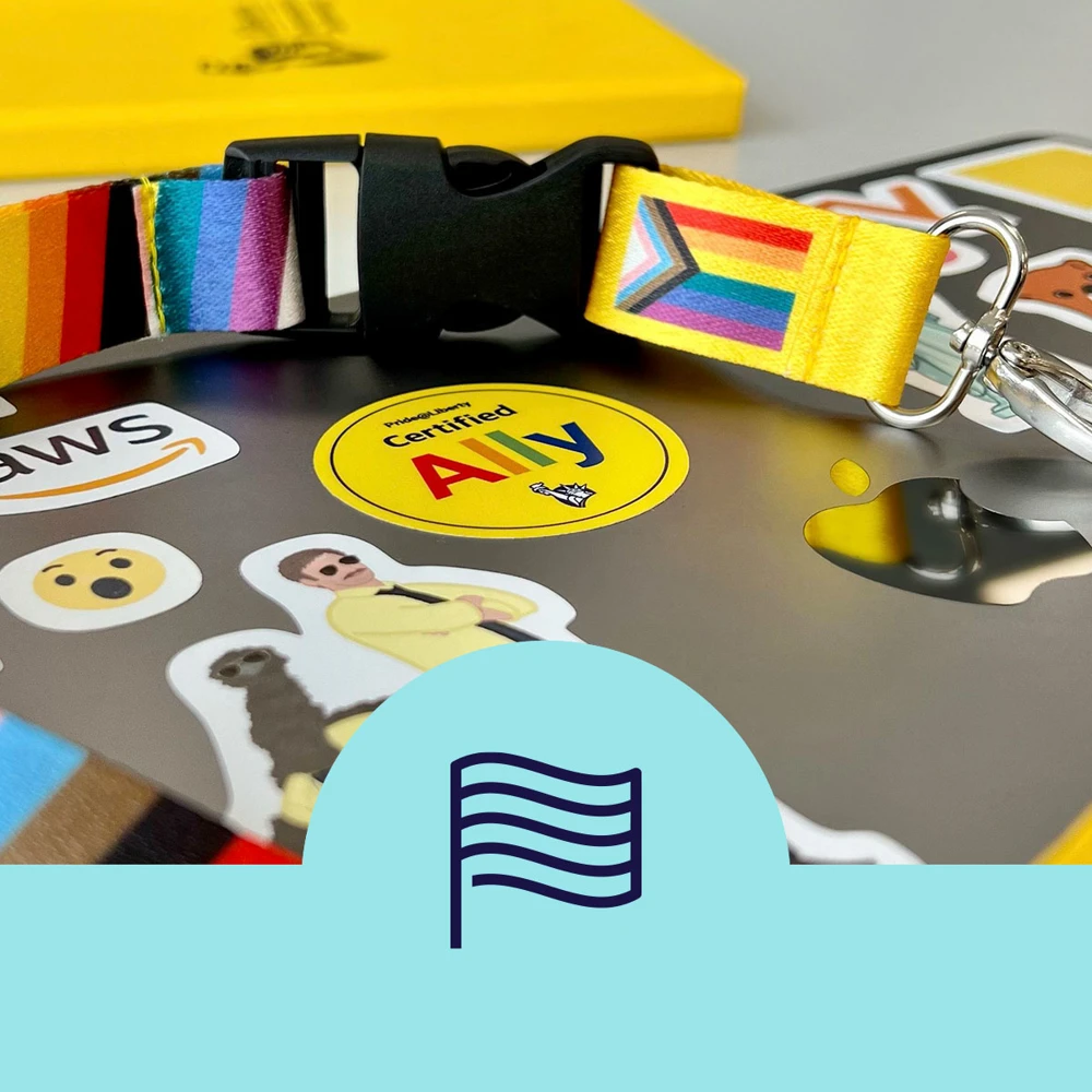 A colorful lanyard with a clip, featuring a rainbow and trans pride design, is laid over a digital device adorned with various stickers, including a "Certified Ally" sticker, an Apple logo, a smiley face, and a graphic of a person. The background includes a bit of yellow and a light blue design with a flag symbol.