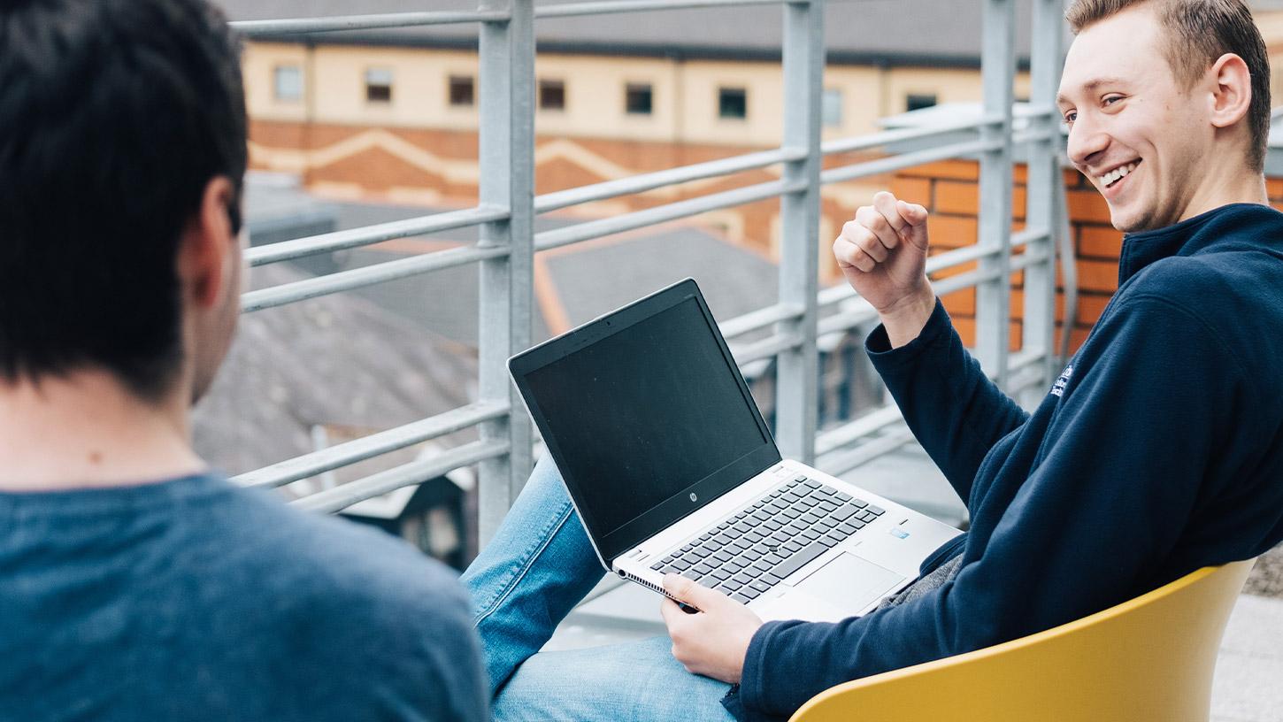 Image of two men sitting on an office balcony with a laptop