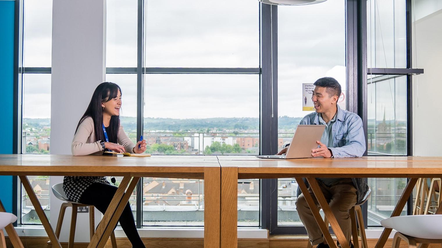 Photo of woman and man sitting at long wooden table, looking at laptop