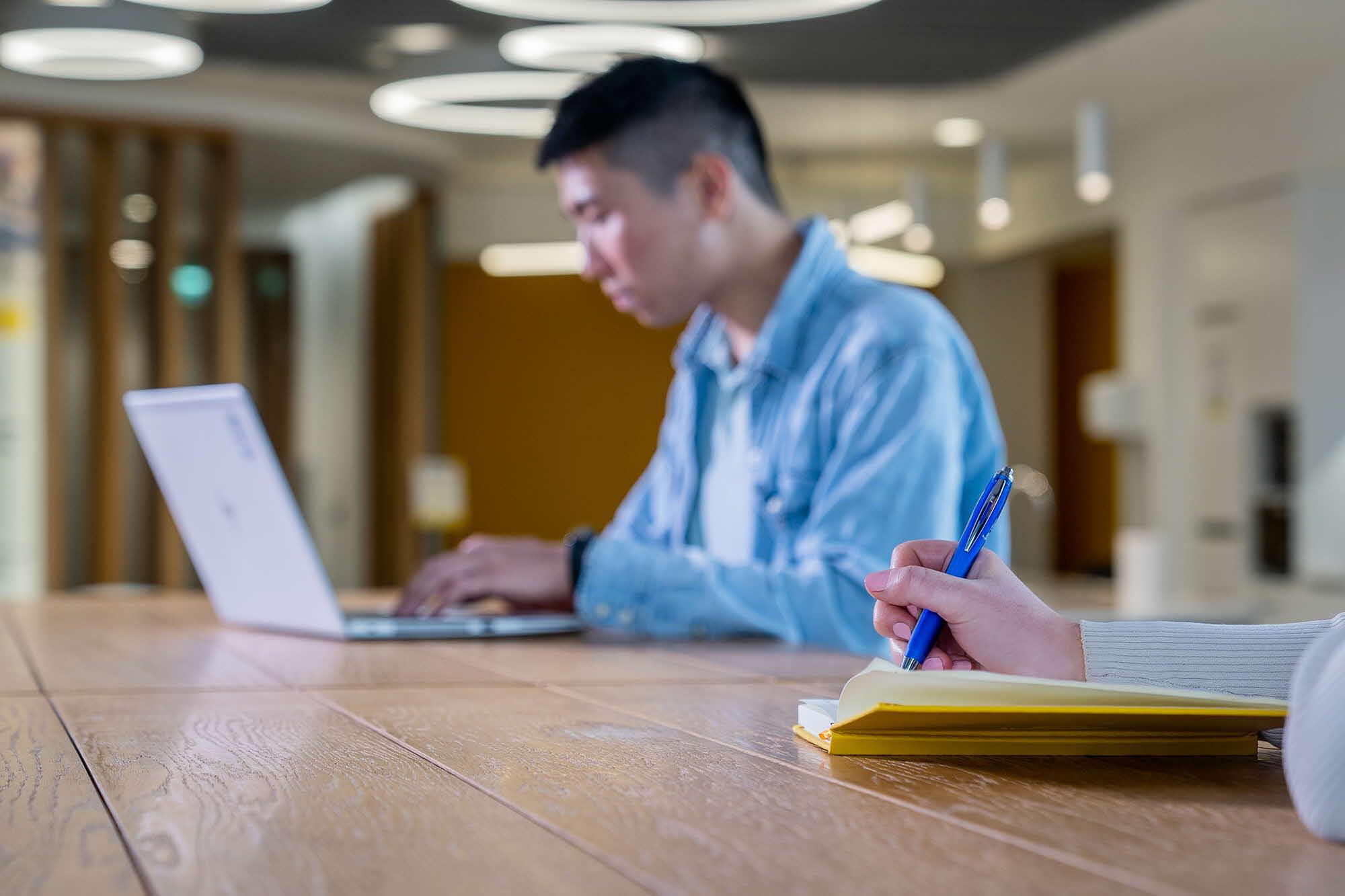 A hand writing in a notebook with a laptop in the distance